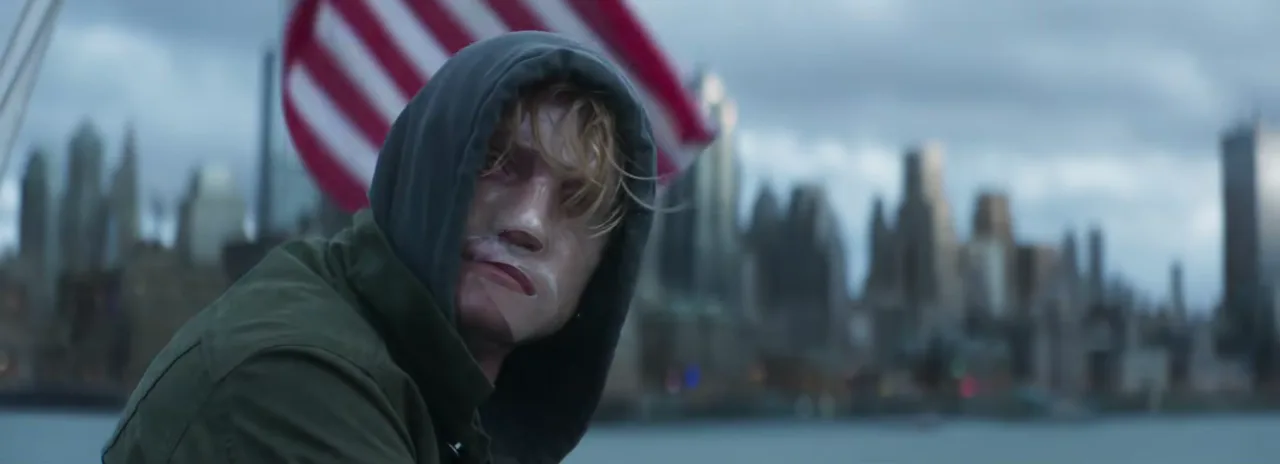 Tom Rhys Harries as Matt Hagen in a green hood, his face scarred and stiff, in front of the Gotham City skyline and an American flag