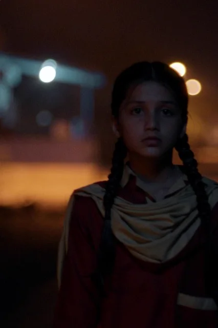 Nazualiya Arsalan as Rabia, in a maroon school uniform and yellow scarf, standing on a village street at night
