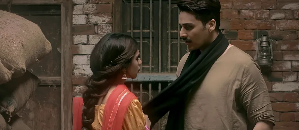 A man in a black shawl facing a young woman in a red dupatta in a brick-walled storeroom, in period costume