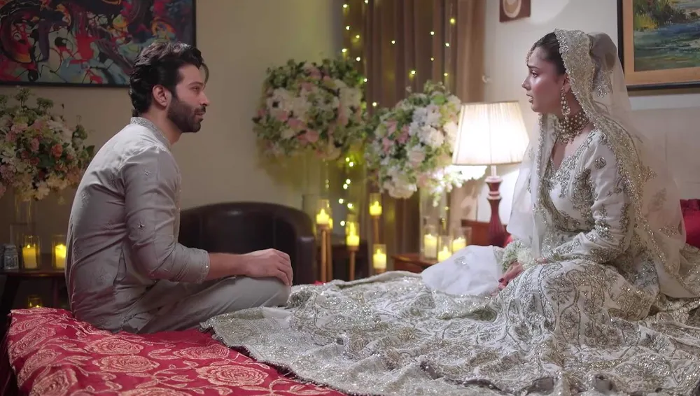 A groom sitting on a red bedspread facing a bride in a silver lehenga, in a room lit with candles and flowers