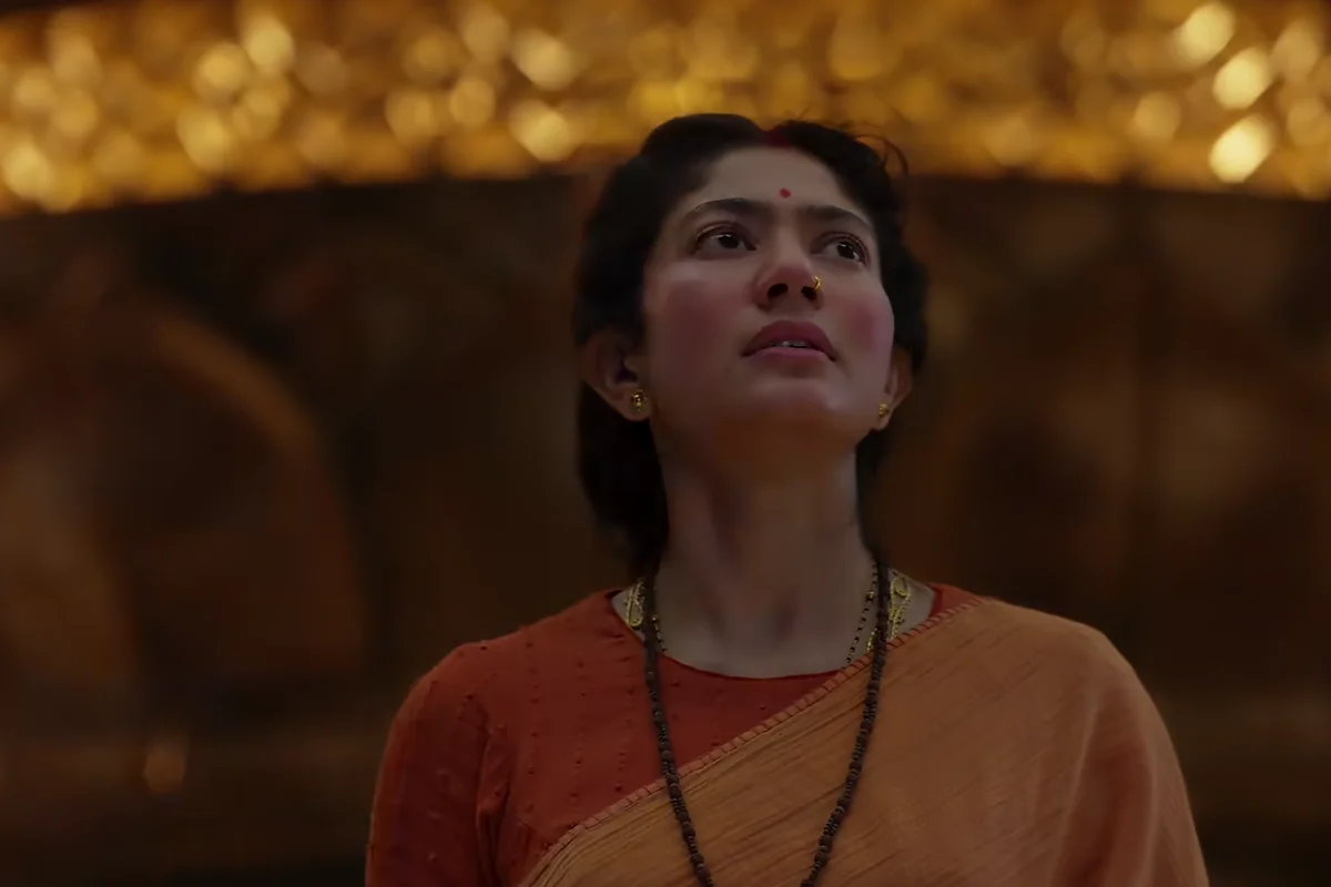 Sai Pallavi as Sita in a saffron sari, looking up in a dimly lit golden hall