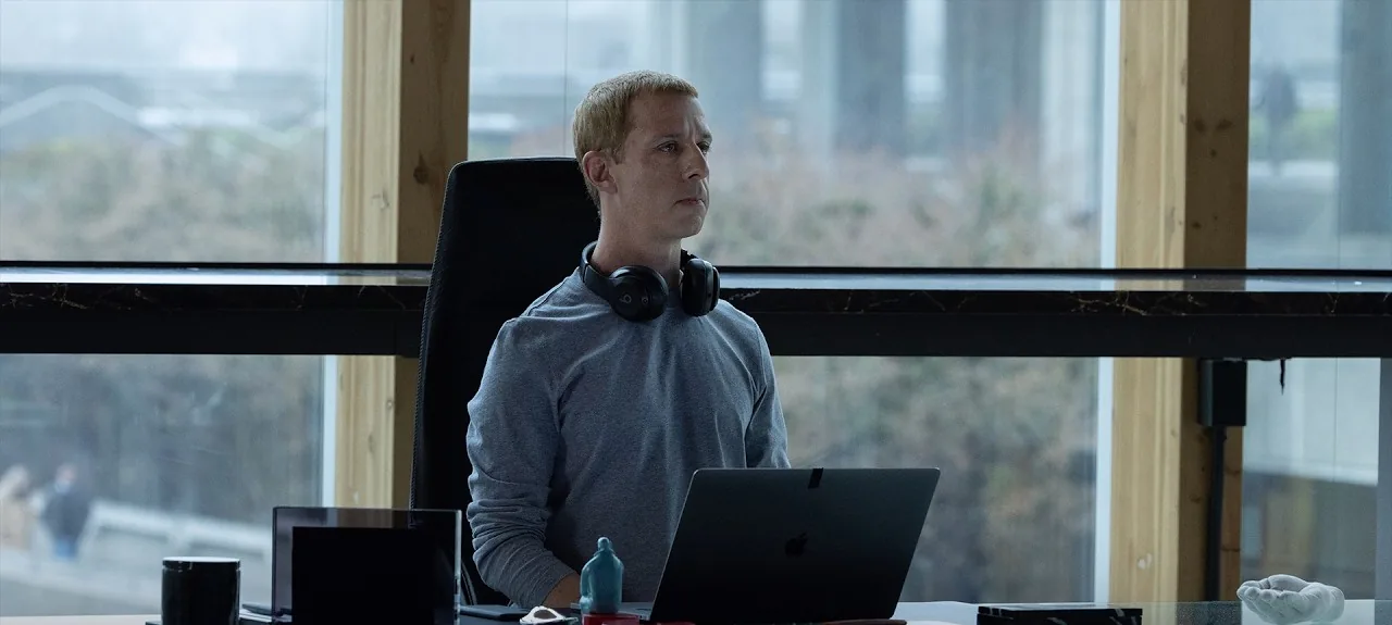 Jeremy Strong as Mark Zuckerberg, headphones around his neck, sitting at a glass desk in an office, from the official trailer for The Social Reckoning